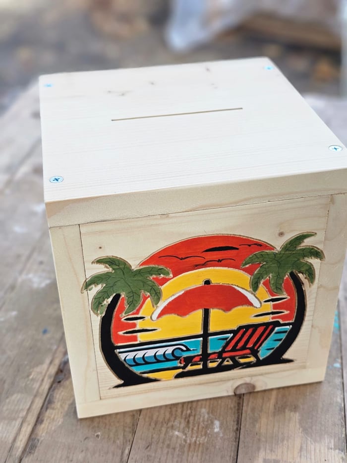 A pine piggy bank with a sunset, palm trees and a beach chair carved and painted on the front
