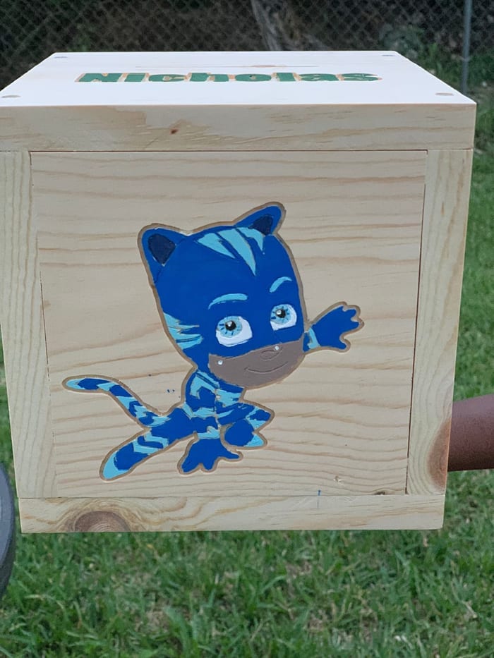 A pine piggy bank with PJ Masks Catboy carved and painted on the front and the name Nicholas on the lid
