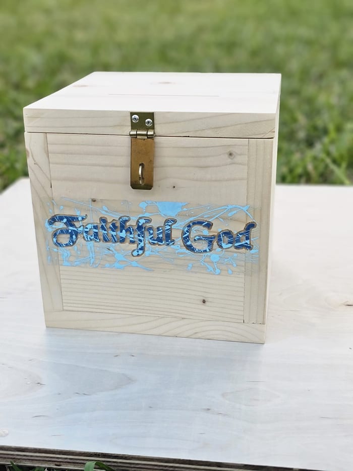 A pine box carved with the words Faithful God in blue over a splattered background