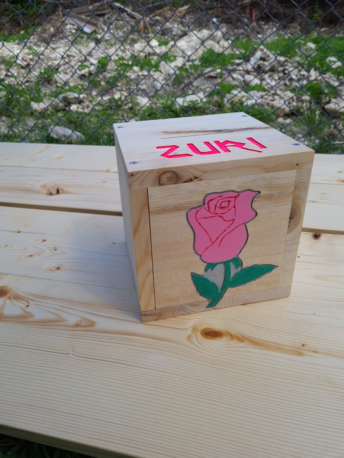 A pine piggy bank with a pink rose carved and painted on the front and the name Zuri on the lid